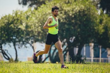 Full length of young bearded sportsman in activewear running on grassy lawn in city park near green trees during outdoor workout