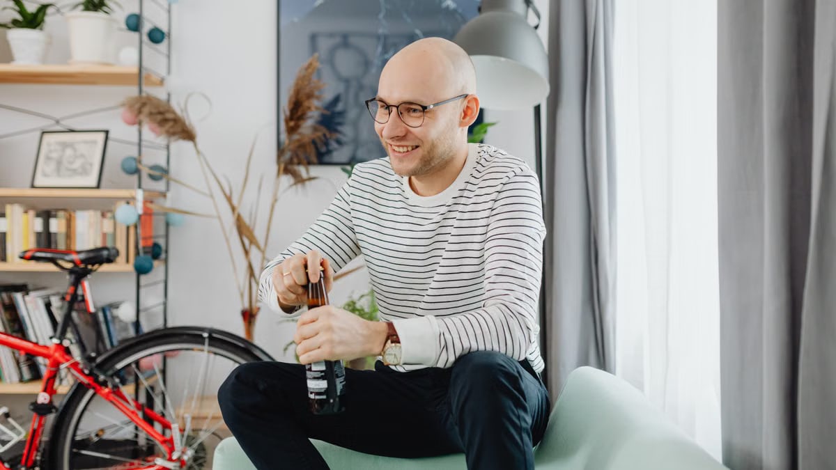 Smiling bald man opening a beer bottle while sitting on a sofa in a stylish living room.